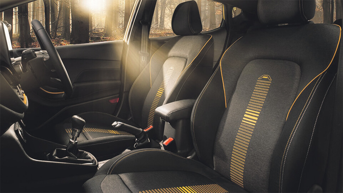 Interior view of a modern car showing black fabric front seats with yellow stitching and decorative yellow stripe patterns, a black steering wheel, manual gear shift, and armrest in soft natural sunlight coming through the windows, highlighting a forest setting outside; suitable for used cars and new cars.
