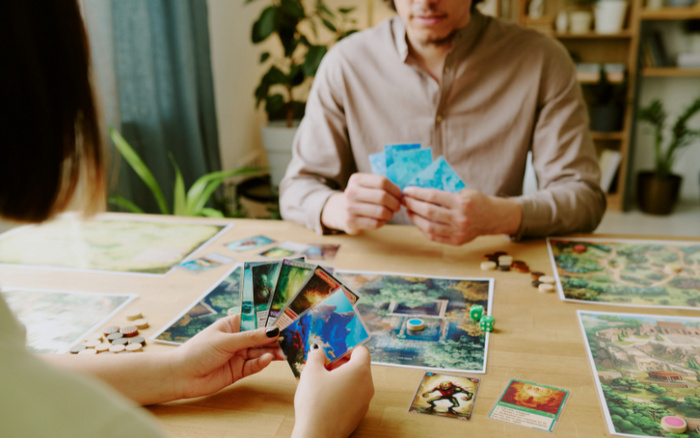 Two people playing a fantasy-themed card and board game at a wooden table with detailed illustrated cards and game boards featuring mystical creatures and landscapes; one person holds a hand of colourful cards while the other is selecting cards, surrounded by game pieces and dice in a cozy indoor setting with plants and shelves in the background. No visible text related to used cars or new cars.