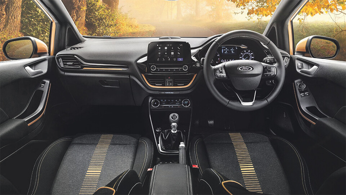 Interior view of a Ford car with a black and grey dashboard and seats featuring gold accents. The steering wheel, displaying the Ford logo, is on the right side, indicating a right-hand drive vehicle. The centre console includes a touchscreen display and manual gear stick. Outside the windows, autumn foliage is visible, creating a warm, natural backdrop. This image relates to used cars and new cars.
