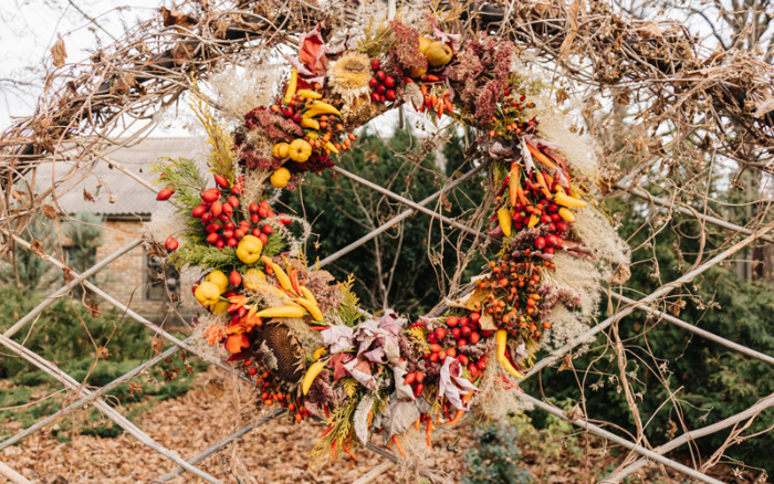 Decorative autumn wreath made of dried flowers, leaves, berries, and small yellow and orange peppers, hanging on a wooden lattice structure outdoors, with a blurred background of trees and fallen leaves, no text present, not related to used cars or new cars