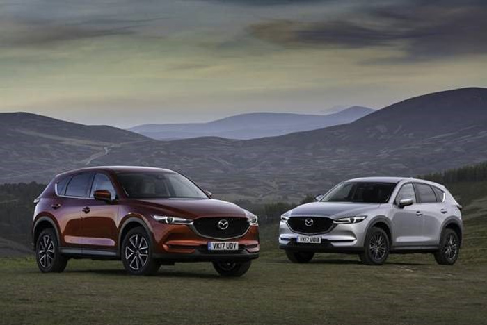 Two Mazda CX-5 SUVs parked on a grassy hill with rolling hills and a moody sky in the background. One car is a metallic brown colour with the license plate VK17 UDV, and the other is silver with the license plate WV17 UDB. The scene highlights the vehicles as options for both used cars and new cars in a scenic outdoor setting.