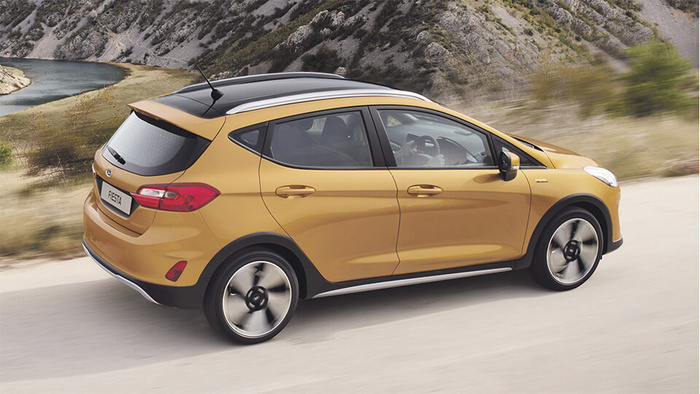 Gold coloured Ford Fiesta hatchback car driving on a road beside a rocky hillside and river, showcasing sporty alloy wheels and black roof with roof rails, representing options for used cars and new cars.