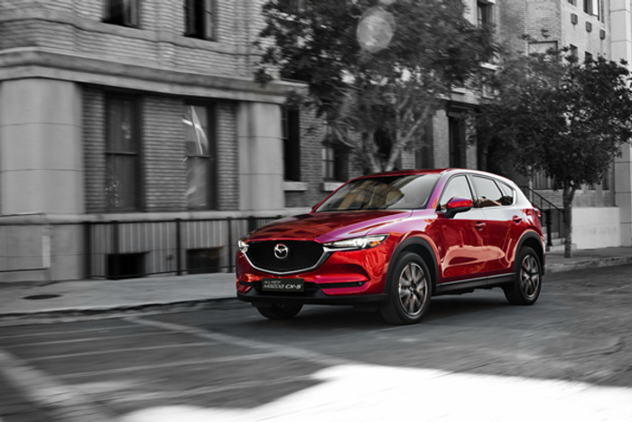 Red Mazda CX-5 driving on an urban street with grey buildings in the background, showcasing a sleek design. Image relevant for used cars and new cars listings.