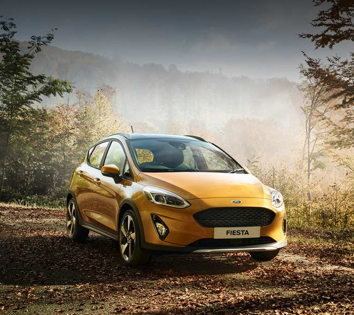 Golden yellow Ford Fiesta parked on a forest path covered with fallen leaves, surrounded by trees with autumn foliage and mist in the background, representing a new car model suitable for both used cars and new cars buyers.