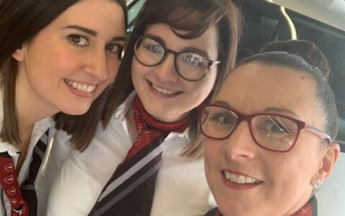 Three smiling women wearing white shirts, glasses, and red scarves with a pattern, posing for a close-up photo inside a car, representing staff at a dealership offering used cars and new cars.