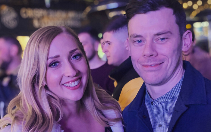 Smiling young woman and man in a social setting with blurred background featuring indistinct text, no visible signs related to used cars or new cars.
