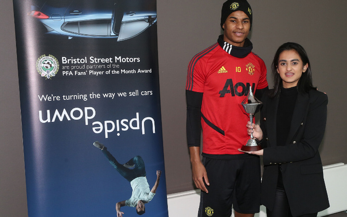 A man in a red Manchester United football kit and black beanie hat, and a woman in a black blazer holding a trophy together. Next to them is a blue banner for Bristol Street Motors, partners of the PFA Fans' Player of the Month Award, with the slogan 'We're turning the way we sell cars upside down' and an image of an upside-down footballer. The banner promotes used cars and new cars.