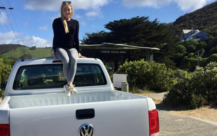 A woman in a black sweater and grey leggings sitting on the back of a white Volkswagen pickup truck parked near a scenic area with green hills and trees. In the background, a wooden sign reads 'GREAT OCEAN ROAD'. The image suggests a lifestyle or travel theme relevant to used cars and new cars, highlighting outdoor adventure and vehicle versatility.