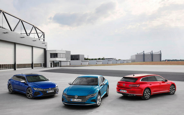 Three Volkswagen cars parked on a large concrete area in front of an industrial building. The vehicles include a blue sporty station wagon on the left, a blue sleek sedan in the centre, and a red sporty estate car on the right. The cars have modern design features with sharp lines and LED headlights. The background features a large warehouse with metal framework and an expansive sky with scattered clouds. This image represents a range of new cars and used cars available from Volkswagen.