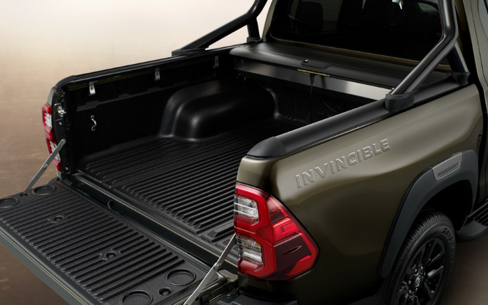 Open rear loading area of a dark green pickup truck with the word INVINCIBLE embossed on the side above the rear wheel. The truck features a ribbed, durable cargo bed liner and a tailgate with metal supports. Rear lights are visible, and the vehicle is presented in a clean, well-lit setting, suitable for advertising used cars or new cars.