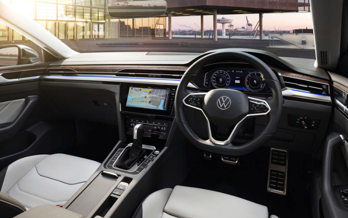 Interior view of a Volkswagen car showing a modern dashboard with a digital display behind the steering wheel, a central touchscreen displaying a navigation map, and light grey leather seats. The steering wheel has the Volkswagen logo. The car is parked in a modern building area with glass walls and reflections visible outside. This image relates to used cars and new cars.