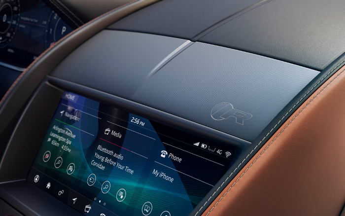 Close-up of a car dashboard featuring a digital infotainment screen displaying navigation to Arlington Avenue, Leamington Spa, weather information, and music playing 'Young Before Your Time' by Convidae via Bluetooth audio. The screen also shows phone connectivity with My iPhone paired. The top of the dashboard has a textured panel with an 'R' logo, and the surrounding interior features brown leather upholstery. The image highlights modern car technology in both used cars and new cars.