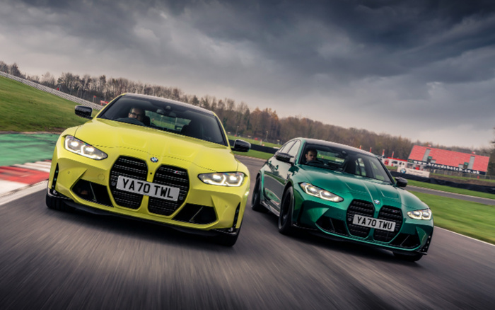 Two high-performance BMW cars driving side by side on a race track under a cloudy sky. The car on the left is a yellow BMW with license plate YA70 TWL, and the car on the right is a green BMW with license plate YA70 TWU. Both cars feature prominent kidney grilles and aggressive front designs, representing new cars in sporty conditions. The scene evokes the dynamic appeal of new and used cars for driving enthusiasts.
