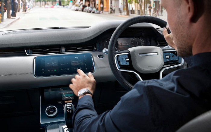 Interior view of a modern car dashboard featuring a man operating the central touchscreen display. The touchscreen shows media controls with options like audio, phone, and navigation. The digital speedometer shows 0 mph. The steering wheel has multiple control buttons and a digital display behind it. The scene is set on an urban street visible through the windshield, illustrating a modern vehicle's user interface, relevant for used cars and new cars searches.