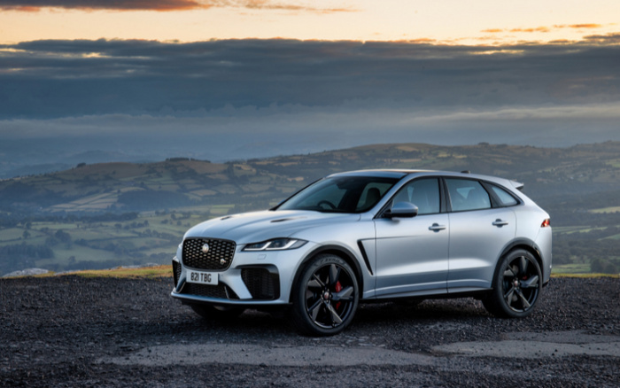 Silver Jaguar F-PACE SUV parked on a gravel surface with rolling hills and a cloudy sky at sunset in the background. The car has a visible UK registration plate reading B21 TBG, black alloy wheels with red brake calipers, and sleek modern design. Ideal image representing used cars and new cars in scenic countryside setting.