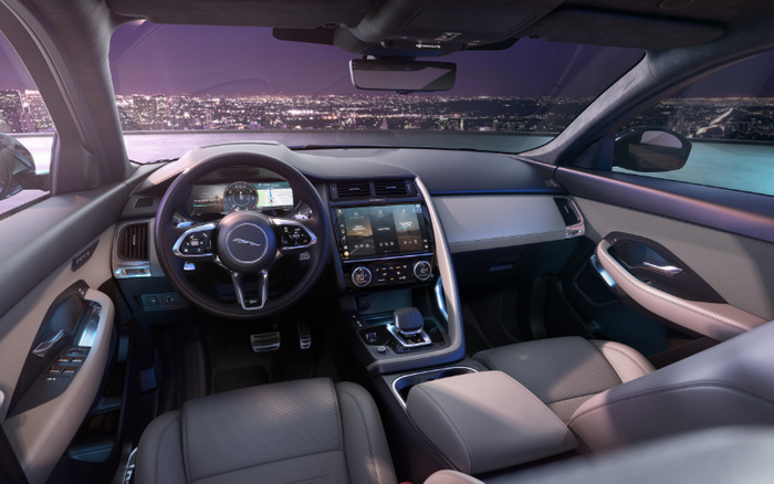 Interior view of a modern Jaguar car showing the steering wheel with the Jaguar logo, digital instrument cluster, large central touchscreen display with climate control settings, beige leather seats, and door panels. City skyline with lights is visible through the windshield and windows. Ideal for advertising used cars and new cars with luxury and advanced technology features.