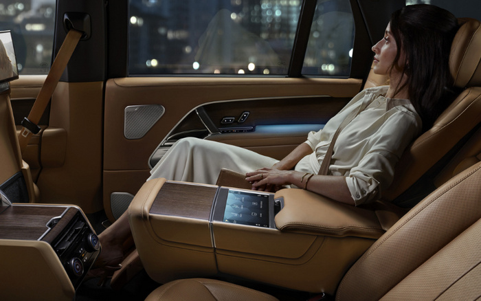 Interior view of a luxury car featuring tan leather seats and wood trim, with a woman seated comfortably in the spacious rear seat looking out the window. The centre console includes a touchscreen panel with various control icons. The car window shows a blurred cityscape at night. The image highlights the premium interior design and comfort of new cars, suitable for promotions involving used cars and new cars.