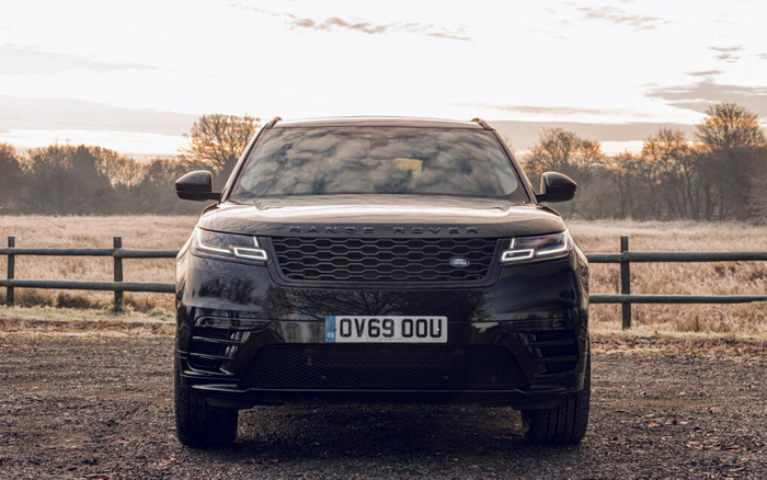 Front view of a black Range Rover SUV parked outdoors on a gravel surface with a wooden fence and leafless trees in the background during early morning or late afternoon. The car has a GB license plate with registration OV69 OOU, illuminated modern LED headlights, and a honeycomb grille. The image is suitable for used cars and new cars advertisements.