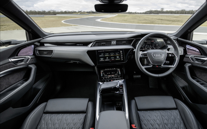 Interior view of a modern Audi car showcasing the driver's seat, steering wheel with Audi logo, digital dashboard displaying speed and navigation, and two central touch screens with vehicle controls. The interior features black leather seats with diamond stitching, sleek black and gray dashboard design, and panoramic view of an empty race track outside. Keywords used cars, new cars.