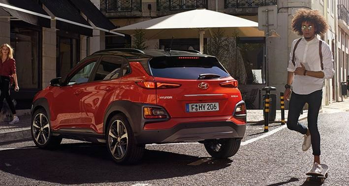 Red Hyundai Kona parked on a city street beside a café with outdoor seating under a large white umbrella, showing its rear view with license plate F•HY 206. A young man with curly hair, wearing a white shirt, black trousers with suspenders, and sunglasses is skateboarding nearby. The scene highlights new cars in an urban setting, suitable for promoting used cars and new cars.