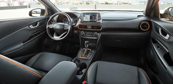 Interior view of a modern car featuring a Hyundai logo on the steering wheel, digital infotainment screen displaying navigation, and a central console with gear shift lever. The car has black seats with orange stitching, air vents with orange accents, and buttons for media and climate control. This image represents the interior quality of used cars and new cars.