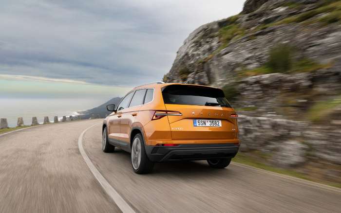 Orange Škoda Karoq driving on a winding coastal road next to rocky cliffs, showing the rear view with European license plate 5SM 3582, representing used cars and new cars options.