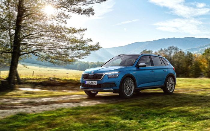 Blue Škoda SUV driving on a dirt path through a grassy field with trees and mountains in the background under a partly cloudy sky, illustrating a new car model suitable for both off-road and city driving, ideal for customers looking for used cars or new cars.
