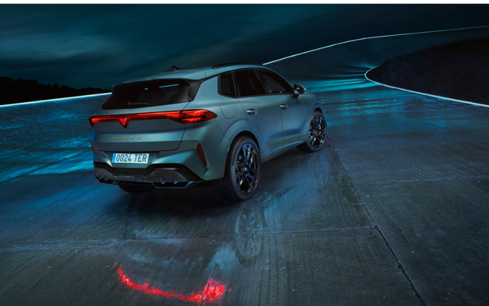 Rear view of a matte green CUPRA Formentor SUV parked on a night-time concrete surface with illuminated blue lines in the background. The vehicle has dark alloy wheels, a license plate reading '0024 TER,' and distinctive red LED rear lights reflecting on the ground. Ideal for showcasing used cars or new cars in a modern setting.