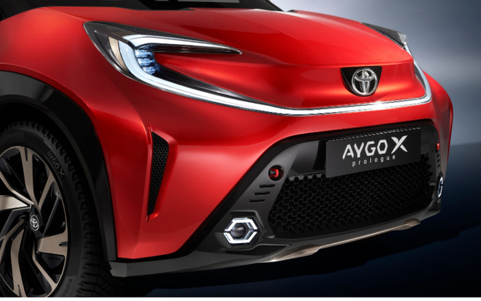 Close-up front view of a red Toyota Aygo X Prologue car highlighting the LED headlight, Toyota logo on the bonnet, distinctive front grille with hexagonal fog light housing, and sleek modern design. Suitable for new cars and used cars buyers looking for a compact city vehicle.