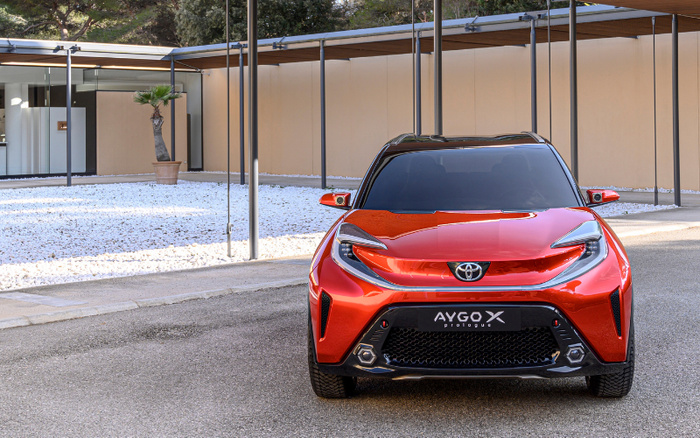 Front view of a shiny red Toyota Aygo X Prologue car parked on a driveway in front of a modern building with beige walls and gravel landscaping. The car features sleek LED headlights, a prominent Toyota badge, and a black grille area with the model name 'AYGO X prologue' clearly visible. Ideal image representing new cars and used cars in a contemporary setting.