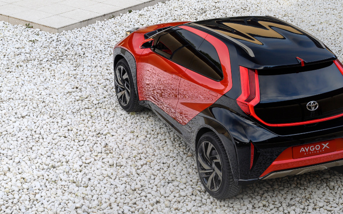 Rear and side view of a red and black Toyota AYGO X Prologue parked on white gravel next to a tiled patio, showcasing modern design and distinctive taillights, suitable for used cars and new cars buyers.