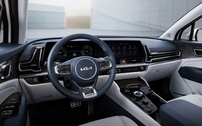 Interior of a modern Kia car showing the steering wheel with the Kia logo, digital instrument cluster, and a widescreen infotainment display with various icons. The dashboard has sleek black and light grey accents, touch controls for climate settings, and a futuristic gear selector. The image highlights the luxurious and high-tech cabin design suitable for both used cars and new cars.