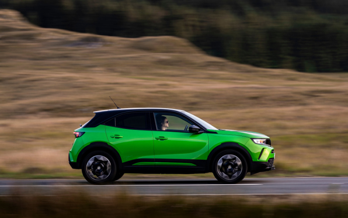 Green compact new car driving on a countryside road with blurred background of dry grassy hills; black roof and modern design, motion blur showing speed; suitable for used cars and new cars vehicle advertising.