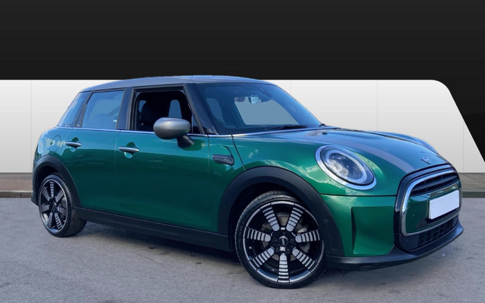 Green Mini Cooper 5-door hatchback parked on a grey surface with modern white panelled backdrop, showcasing sleek black and silver alloy wheels, circular LED headlights, black side mirrors, and tinted windows, suitable for used cars and new cars market