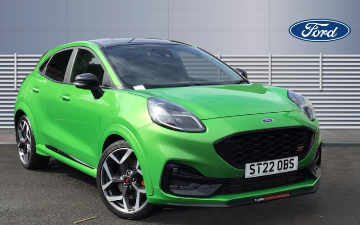 Green Ford Puma ST parked on asphalt with metal fence in the background, UK registration plate ST22 OBS, Ford logo in the top right corner, front grille with honeycomb pattern, 'Ford Performance' text on the front splitter, visible alloy wheels with red brake calipers, suitable for used cars and new cars listings.