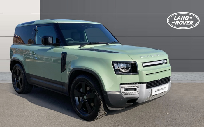 Green Land Rover Defender SUV parked on a paved surface in front of a grey building wall. The vehicle has black tinted windows and black alloy wheels. The front grille displays the Land Rover badge with Defender lettering on the bonnet. A white plate on the front bumper reads 'APPROVED'. The Land Rover logo is visible on the upper right corner of the image. Ideal for used cars or new cars listings featuring Land Rover vehicles.