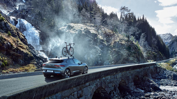 Silver Jaguar I-PACE car with a roof-mounted racing bicycle driving on a stone bridge in a mountainous area with trees and a waterfall in the background. The car's visible license plate reads '171 UYP'. This scenic image highlights the Jaguar I-PACE, representing new cars suitable for active lifestyles, relevant for buyers of used cars and new cars.