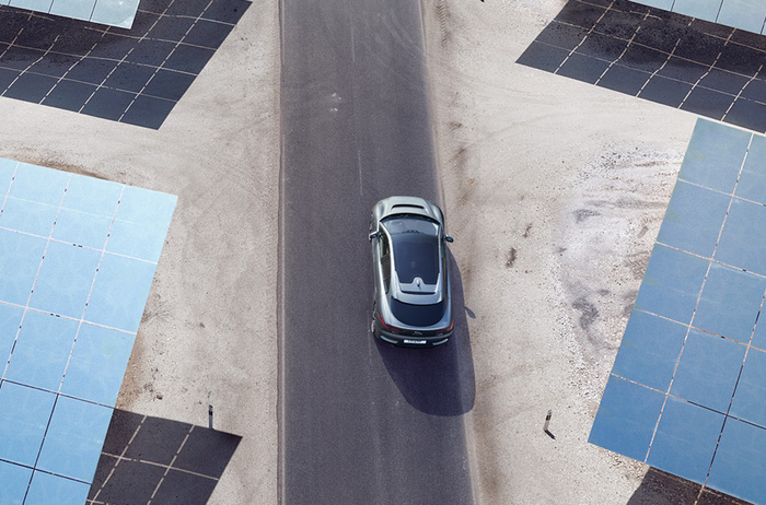 Aerial view of a silver car driving on a paved road surrounded by large blue solar panels on sandy ground, representing a clean energy environment suitable for both used cars and new cars.