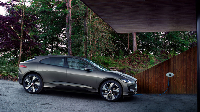 Grey Jaguar electric car parked and charging under a modern carport with wooden panel walls and surrounded by green forest trees, representing new cars and used cars in eco-friendly automotive technology.