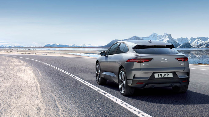 Rear view of a silver Jaguar I-PACE EV400 electric vehicle driving on a coastal road with a scenic backdrop of snow-capped mountains and reflective water under a clear blue sky, licence plate 171 UYP, showcasing a modern new car ideal for used cars and new cars buyers.
