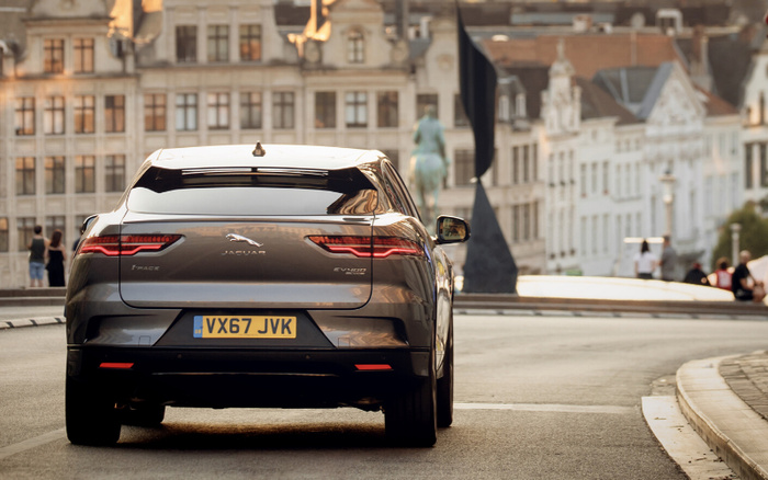 Rear view of a grey Jaguar I-PACE EV400 electric car with registration plate VX67 JVK driving on a curved urban street, showcasing sleek taillights and modern design, suitable for illustrating used cars and new cars in city settings.