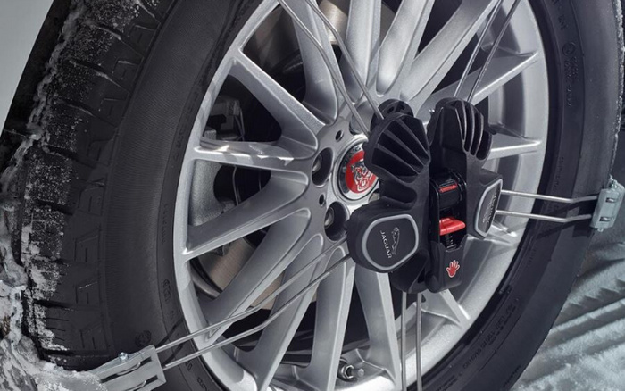 Close-up of a Jaguar car wheel with a tyre traction device fitted. The silver alloy wheel features the Jaguar logo at the centre. The black traction device is attached with metal wires, designed to improve grip on snowy or icy roads. Snow is visible on parts of the tyre and ground, highlighting winter conditions. This image can represent maintenance and safety for both used cars and new cars.