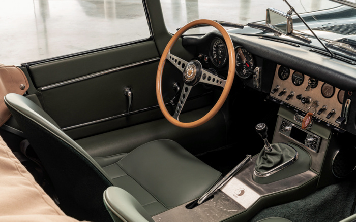 Interior of a classic car featuring a wooden steering wheel with a Jaguar emblem, green leather seats, and a manual gearshift. The dashboard showcases multiple circular gauges and switches, highlighting vintage styling. The image represents the interior of a well-maintained used car, illustrating classic automotive design.