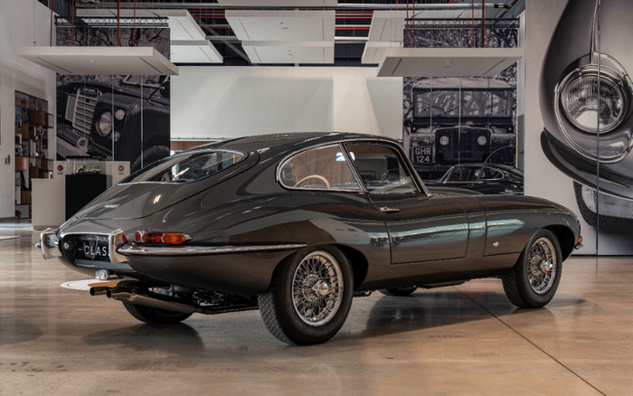 Classic dark grey Jaguar E-Type sports car displayed indoors with rear and side view visible, set against large black and white images of vintage car front grills and headlights on the walls, emphasising a showroom environment for used cars and new cars.