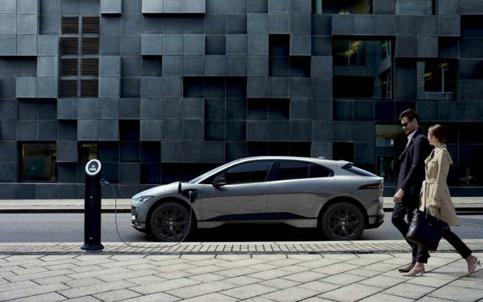 Grey electric car charging at a street charging station with a modern dark tiled building in the background. A man and woman dressed in formal attire are walking past the car on the sidewalk. The scene represents contemporary urban transportation, suitable for illustrating used cars and new cars.