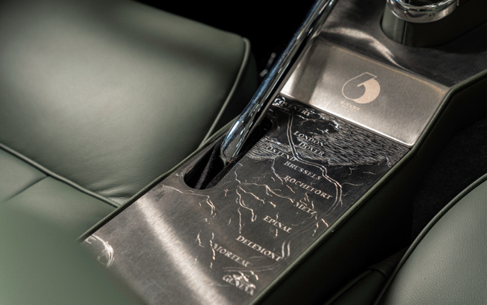 Close-up of a car's centre console featuring a detailed metal plate with engraved names of cities and regions including London, Dover, Brussels, Rochefort, Lille, Edinal, Deinze, Mortien, and Ghent, surrounded by green leather seats, highlighting a luxury car interior design. The image is relevant for used cars and new cars showcasing unique interior craftsmanship.