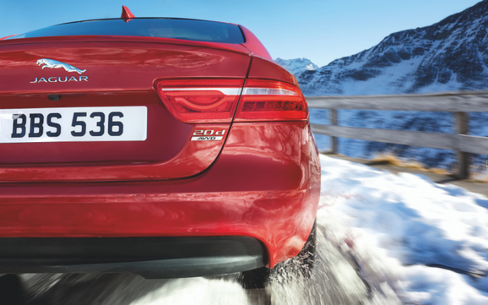 Rear view of a red Jaguar 20d AWD car driving on a snowy mountain road with a wooden guardrail and snow-covered mountains in the background. The car's licence plate reads BBS 536. Ideal image for used cars and new cars related content.