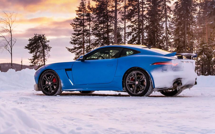 Blue sports car parked on snow-covered ground with rear covered in snow, surrounded by tall pine trees under a colourful sunset sky, representing used cars and new cars in winter conditions.