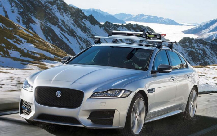 Silver luxury sedan driving on a mountain road with snow-covered peaks in the background, equipped with roof racks carrying skis. Ideal image showcasing new cars in a scenic environment, suitable for advertising new and used cars for winter or adventure driving.