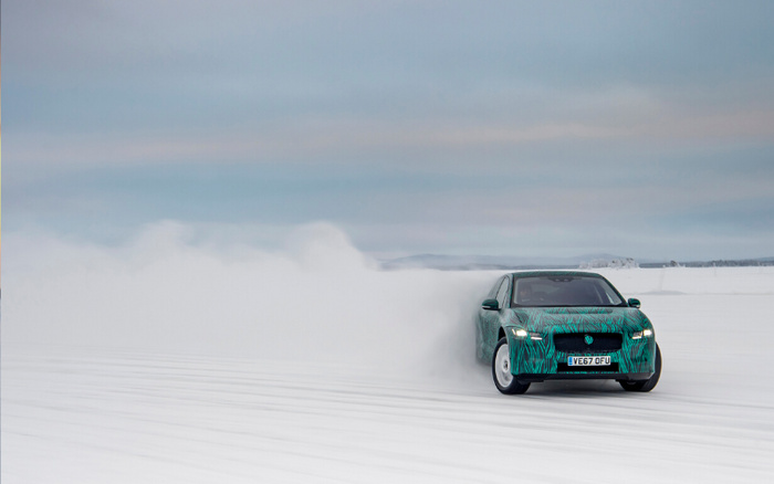 A green and black camouflage Jaguar I-Pace with license plate VE67 OFU drifting on snow, creating a cloud of snow behind it under a cloudy sky, highlighting driving capabilities of used cars and new cars in winter conditions.